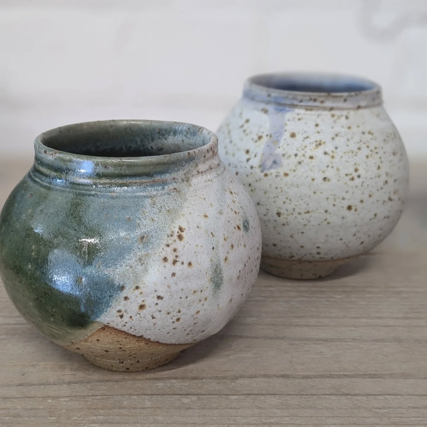 A pair of glazed, ceramic moon pots, on a wooden shelf with a white painted brick wall behind.